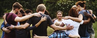 Mehrere junge Menschen stehen im Kreis und fassen sich an den Schultern, die Köpfe gesenkt zum Kreisinneren gewandt. Ein Mädchen blickt zum Betrachter und lächelt.