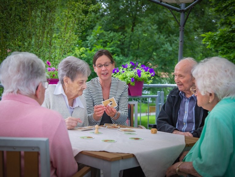 Eine Pflegerin unterstützt Bewohner/innen beim Kartenspiel auf der Terasse