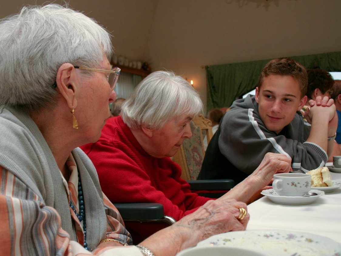 An einem Tisch sitzen mehrere Menschen bei Kaffee und Kuchen zusammen. Ein junger Mann hört einer älteren Dame aufmerksam und mit offenem Blick zu.